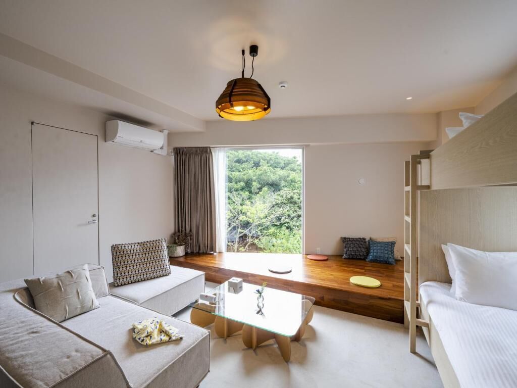 A modern hotel room with a sofa, a small table, a bunk bed, and a large window offering a view of greenery.