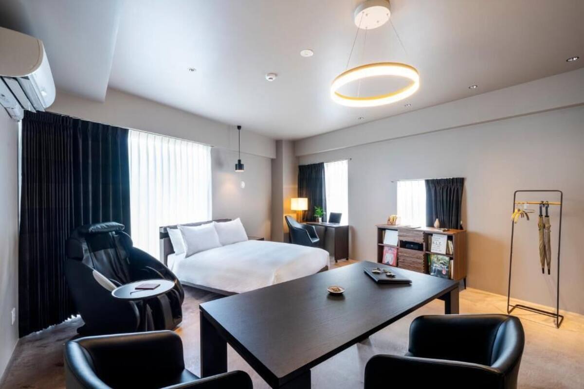 A modern hotel room with a large bed, a desk with a chair, a massage chair, and a standing shower.