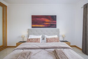 Une chambre à coucher avec un grand lit, deux tables de chevet, une commode, un tableau accroché au mur et une fenêtre avec des rideaux.