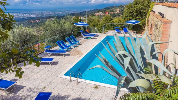 A swimming pool with blue lounge chairs and umbrellas, surrounded by greenery and overlooking a scenic landscape. - Beautiful apartment for 4 guests with WIFI, pool, terrace, pets allowed and panoramic view (Lamporecchio)