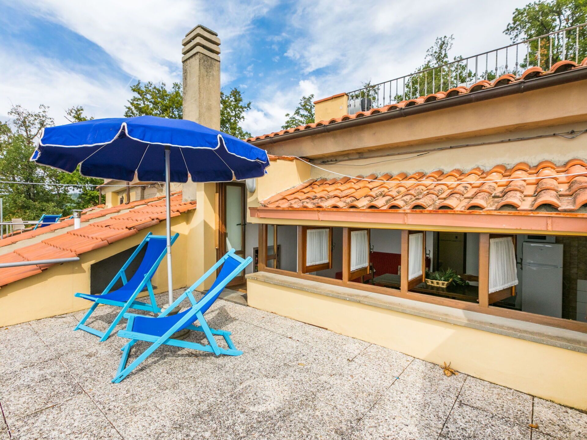 Un bâtiment avec un toit recouvert de tuiles, un balcon avec une chaise bleue et un parasol, et une fenêtre avec des rideaux.