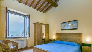 A bedroom with a large bed, a wooden wardrobe, a wicker chair, a window with a view, and a painting on the wall. - Beautiful apartment for 2 guests with pool, WIFI, terrace, pets allowed and panoramic view (Lamporecchio)