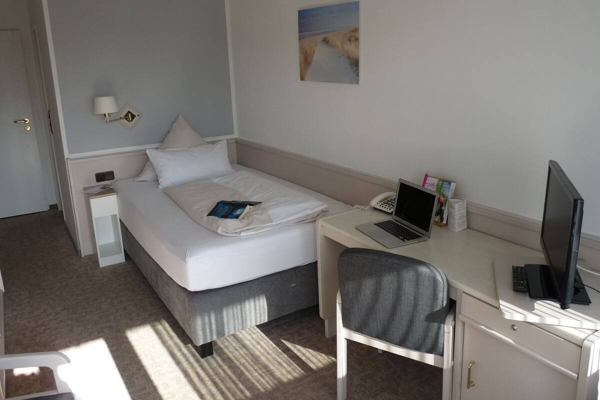 Hotellihuone, jossa on sänky, työpöytä kannettavalla tietokoneella, tuoli, televisio ja taulu seinällä.