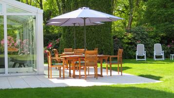 Un patio al aire libre con mesa y sillas, rodeado de abundante vegetación y un cerramiento de vidrio.