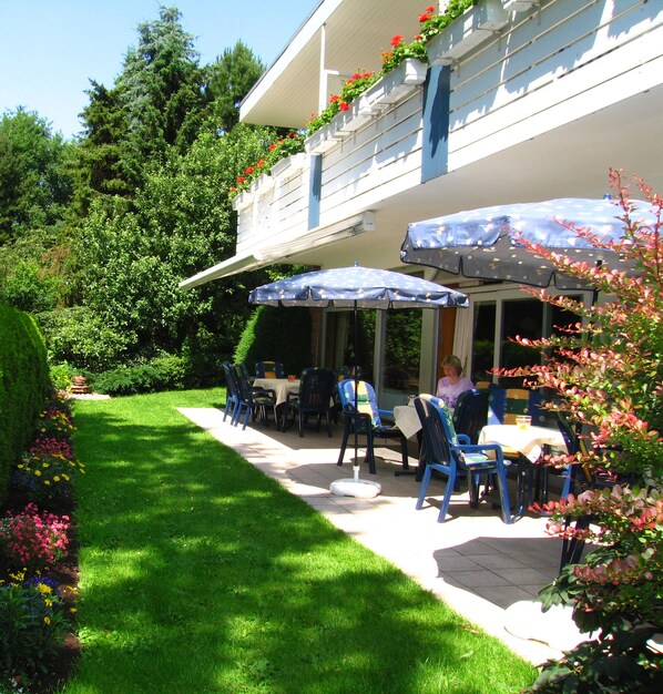 Un patio al aire libre con sillas y sombrillas azules, rodeado de un jardín cuidado y un edificio con balcón.