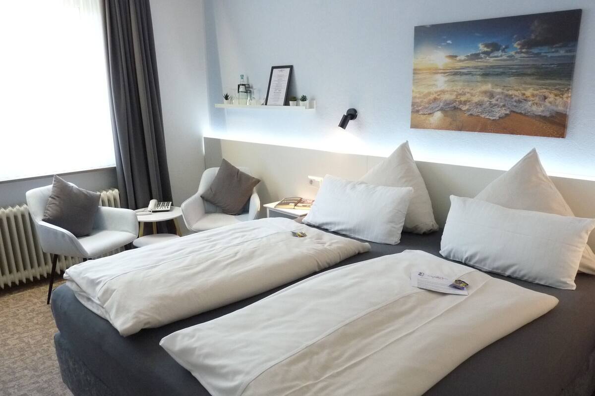 Hotellihuone, jossa on kaksi sänkyä, työpöytä, tuoli, yöpöytä ja seinälle kiinnitetty hylly, jossa on tavaroita.