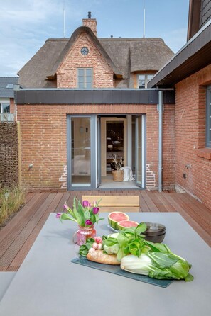 Une maison moderne avec une façade en briques, un toit de chaume et une terrasse en bois agrémentée de légumes frais et de fleurs.