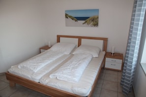 Una cama doble con cabecera de madera, dos mesitas de noche con lámparas, un cuadro colgado en la pared que muestra una playa y una ventana con cortina a rayas.