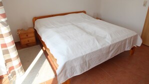 A single-bed room with a wooden frame, a white bedspread, a nightstand, and a wall-mounted light switch.