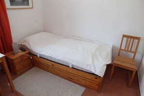 Une petite chambre avec un lit simple, un cadre de lit en bois, une table de chevet, une chaise et un bureau avec une lampe.