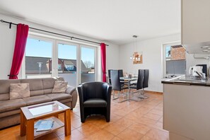 Ett modernt vardagsrum med en soffa, ett matbord med stolar, en köksdel och stora fönster med gardiner.