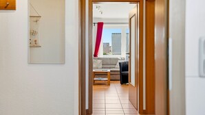 Un couloir mène à un salon où se trouvent un canapé, une table basse et d’où l’on peut admirer la vue sur les bâtiments environnants.