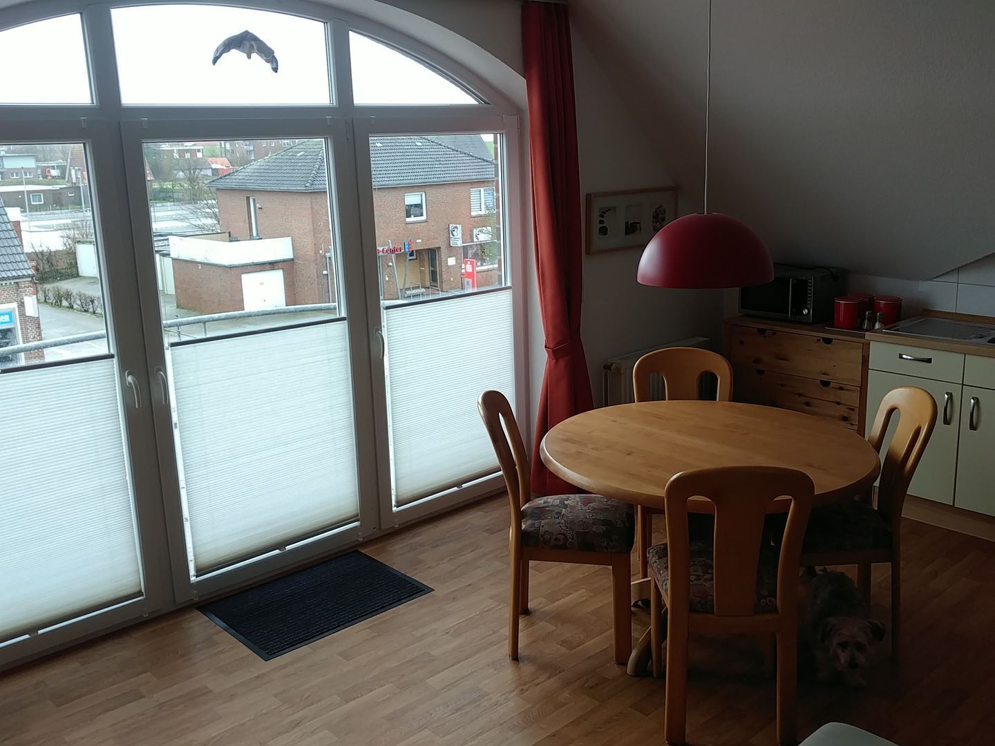 Een kamer met een grote schuifglazen deur, een ronde houten tafel met stoelen, een rode hanglamp en een kleine keuken met kastjes en een magnetron.