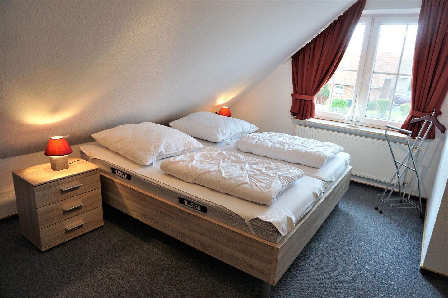 Een slaapkamer met een houten bed, twee nachtkastjes met lampen, een radiator en een raam met gordijnen.