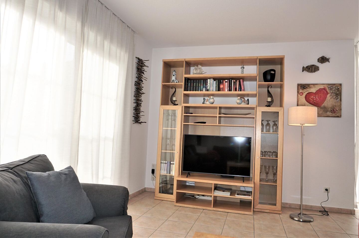 Een woonkamer met een grijze bank, een televisie op een houten dressoir, een vloerlamp en een boekenkast met decoratieve objecten.