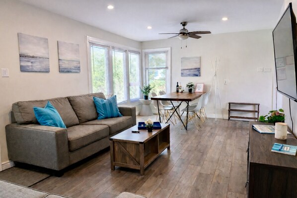 A modern living room with a gray sofa, wooden coffee table, and a dining area with a wooden table and chairs. - Beautiful 3-Bdrm Cottage close to Ski Resorts w/ Hot Tub, Game Room, & more! (Tiny)