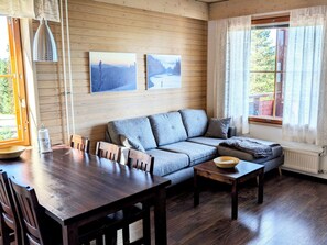Un salon chaleureux avec un plancher en bois, un canapé, une table basse et une table à manger avec des chaises. Deux tableaux encadrés ornent le mur, et une fenêtre est agrémentée de rideaux.