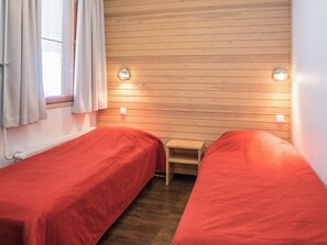 A small room with two single beds, wooden panel walls, a small table, and a window with curtains.