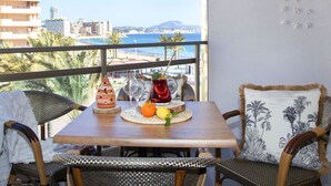 Een balkon met een houten tafel, rieten stoelen en uitzicht op zee en gebouwen.