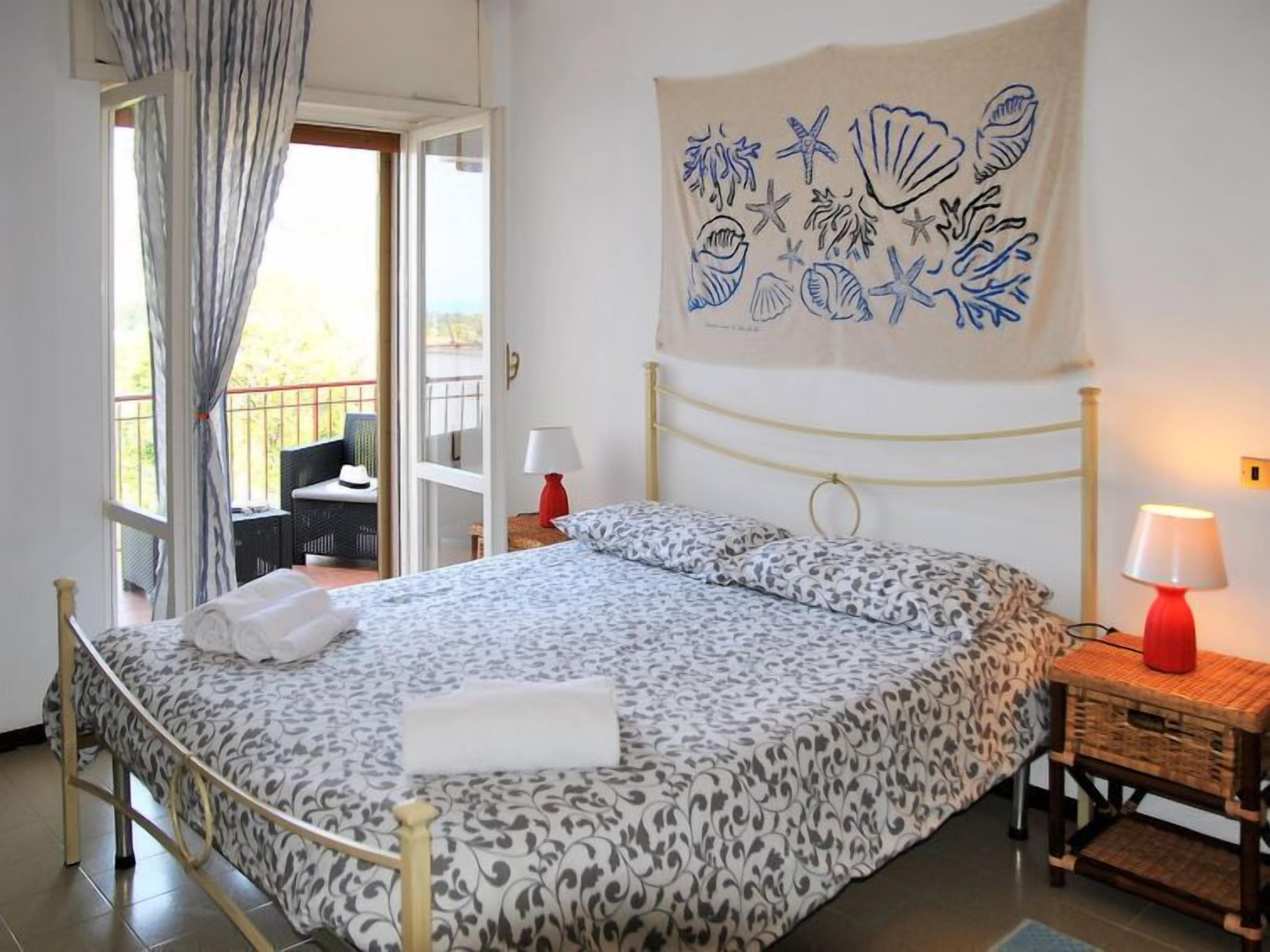 A bedroom with a bed, a nightstand, a lamp, a wall tapestry, and a view of the outside balcony.