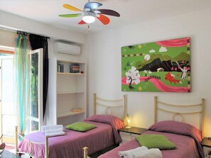 A room with two beds, a ceiling fan, a painting, and a window with curtains.