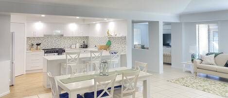 A modern kitchen with white cabinets, a dining area with white chairs and a table, and a living room with a sofa and a TV.