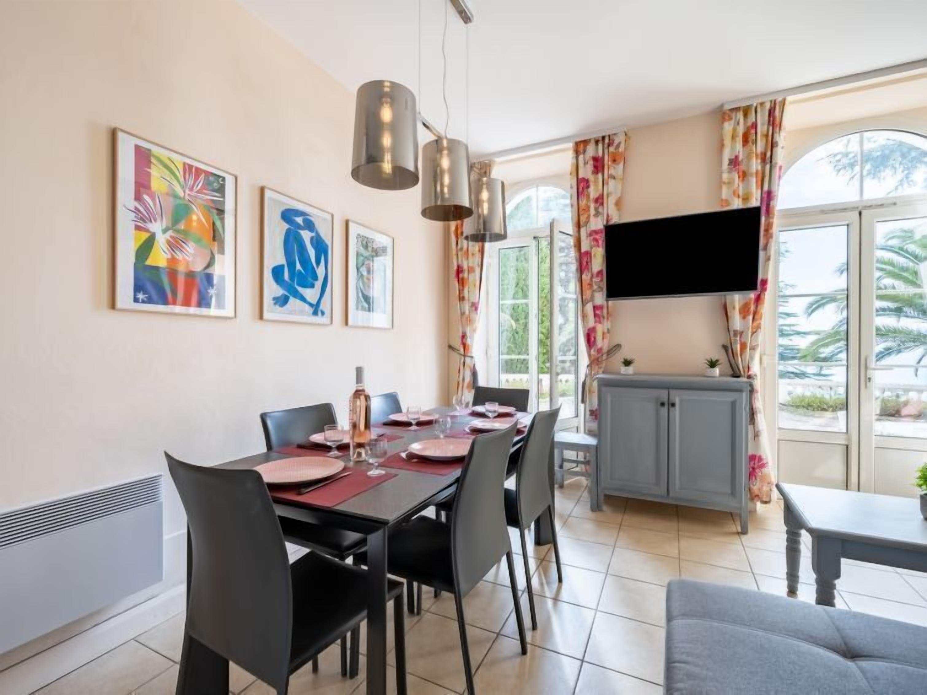 A dining area with a table set for four, chairs, a television mounted on the wall, and a window with curtains.