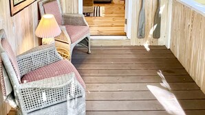 A cozy room with wooden walls, a wicker chair, a lamp, and a view into another room with a couch and a plant. - Charming Folly Cottage! Steps to Downtown, Pier & Beach! (Folly Beach)
