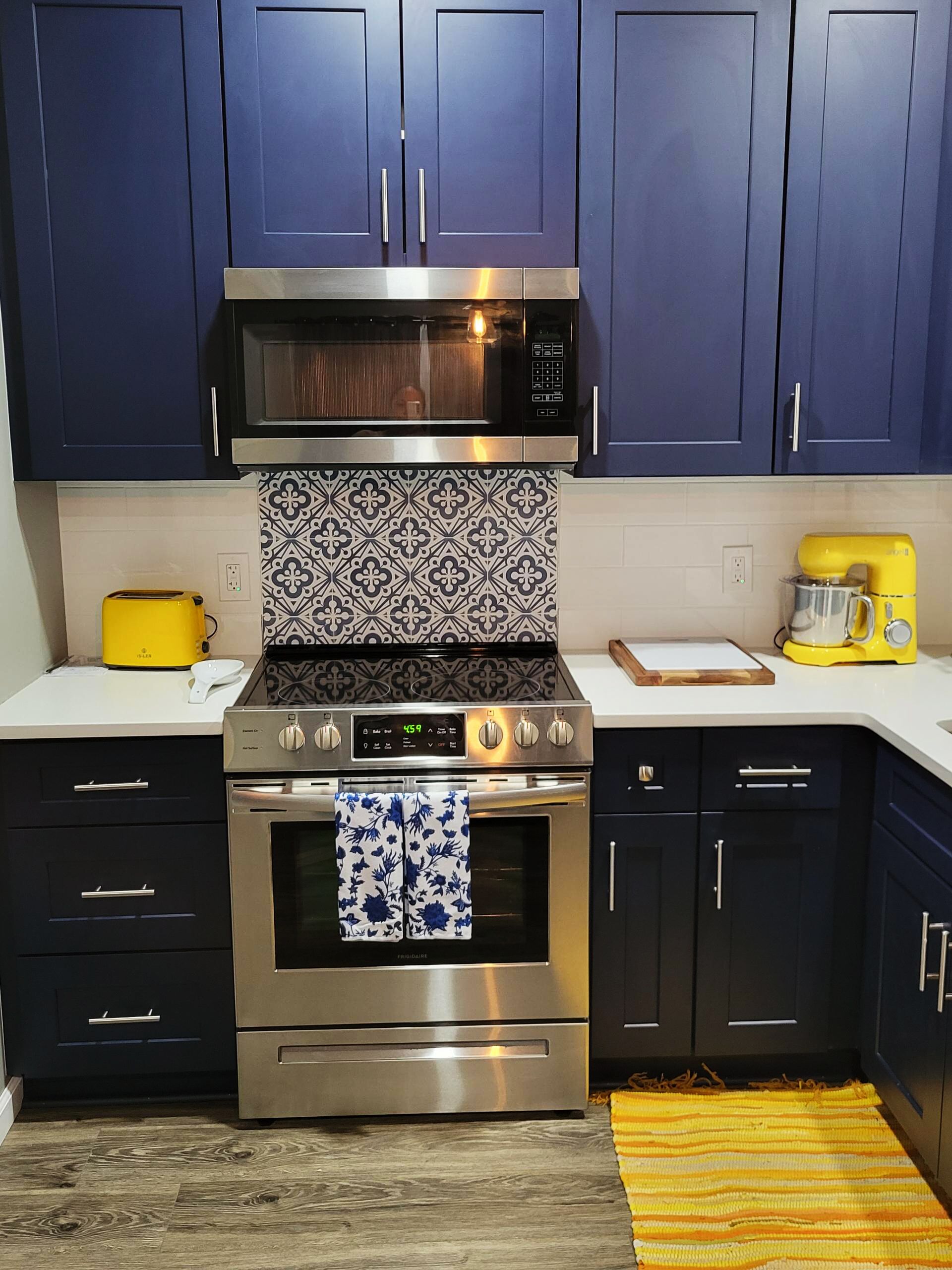 Une cuisine avec des meubles de couleur bleu foncé, une cuisinière en acier inoxydable, un four à micro-ondes, un grille-pain jaune et un revêtement mural à motifs.