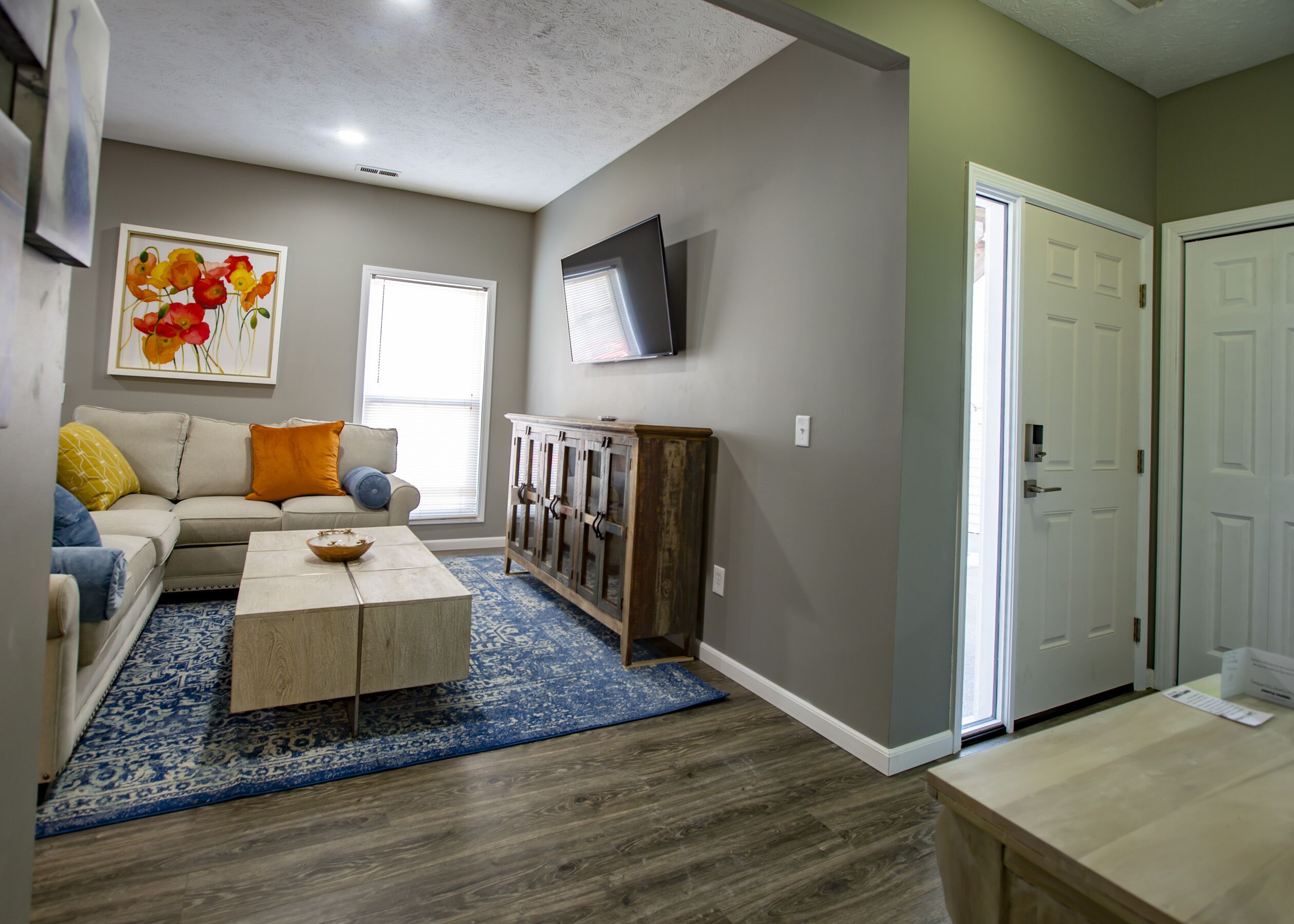 A living room with a sofa, coffee table, and a cabinet. There is a TV mounted on the wall and a door in the background.