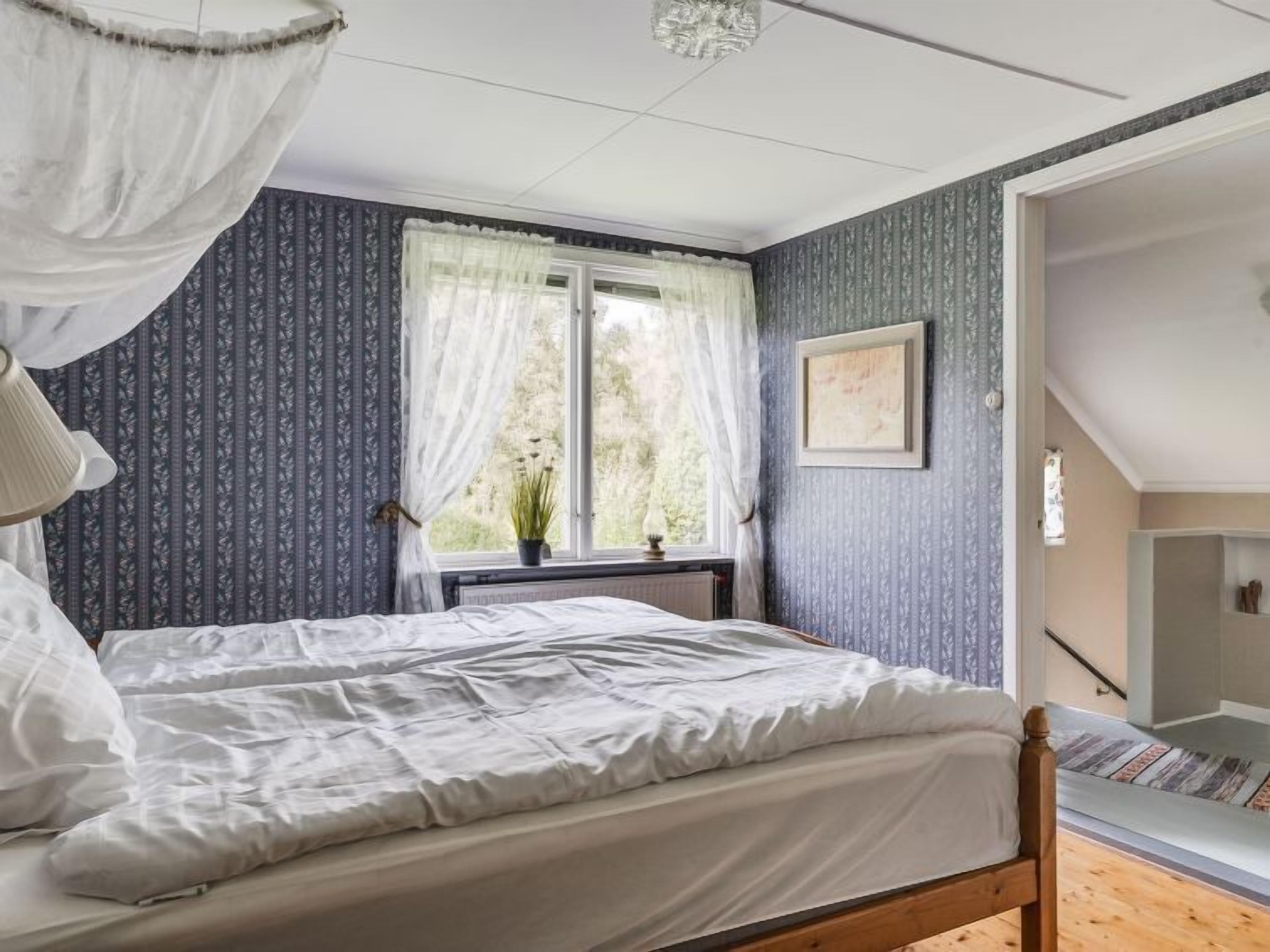 Een slaapkamer met een bed, een raam met gordijnen, een bedlamp en een lijst met een afbeelding aan de muur.