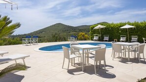 Zona de piscina amb gandules i ombrelles, envoltada de vegetació i vistes a les colines.