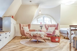 Une chambre à coucher confortable, aménagée en style « loft », avec une grande fenêtre, deux fauteuils roses, un plancher en bois et une armoire blanche.