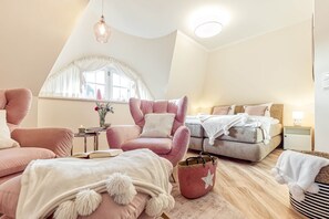 Ein gemütliches Schlafzimmer mit großem Fenster, einem rosa Sessel, einem Bett mit weißer Decke und einem Nachttisch mit Lampe.