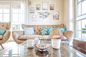 Ein gemütliches Wohnzimmer mit einem beigen Sofa, zwei Sesseln, einem Couchtisch mit Snacks sowie einer Wand mit gerahmten Bildern und einer Vogel-Skulptur.