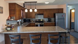 Una cocina con armarios de madera, encimera de granito, refrigerador de acero inoxidable, estufa a gas y tres taburetes de bar.