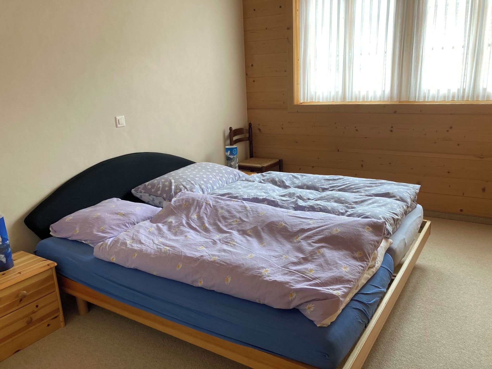 Un lit simple avec un matelas bleu et une literie à motifs floraux, une table de chevet en bois avec des tiroirs, une chaise et une fenêtre avec des stores.