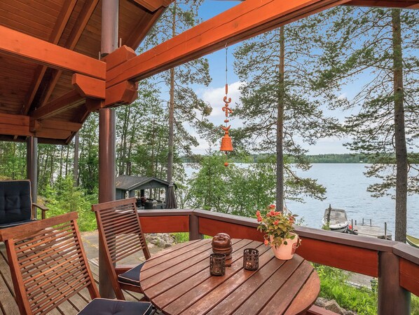 A wooden deck with a table and chairs, overlooking a lake with trees and a dock.