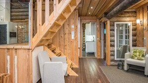 Een houten blokhut van binnen, met een houten trap, een zithoek met een bank en fauteuils, en een keuken zichtbaar door een open deur.
