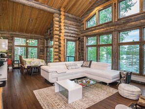 A spacious log cabin interior with a large white sofa, a dining area, and large windows offering views of the forest.