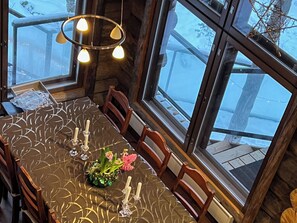 Uma sala de jantar com uma mesa posta para quatro pessoas, cadeiras de madeira, um lustre e janelas amplas com vista para uma paisagem coberta de neve.