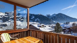 Un balcon avec une balustrade en bois, une table recouverte d’une nappe à motifs floraux et une chaise. On aperçoit, à travers la fenêtre, un paysage montagneux enneigé.