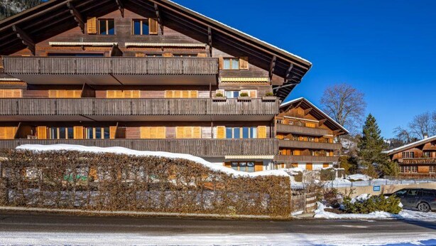 Un chalet en bois à plusieurs étages, doté de balcons, avec un mur de pierre à l’avant et un ciel bleu dégagé.