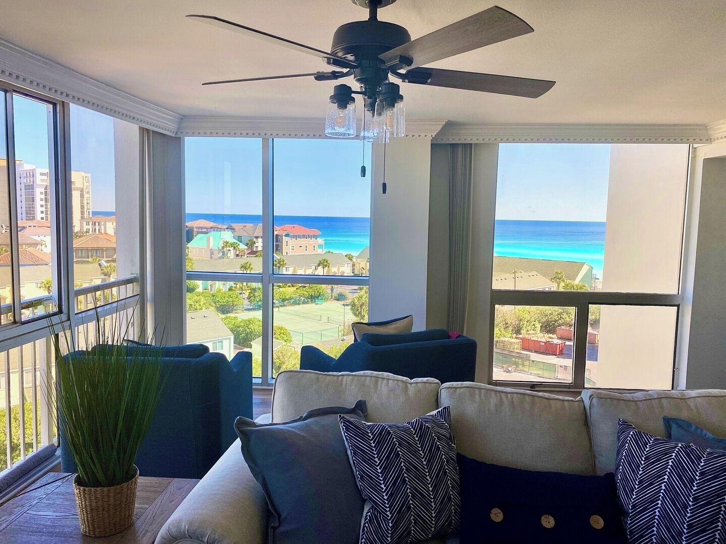 A room with a ceiling fan, large windows with a view of the ocean, a sofa with patterned cushions, and a potted plant.