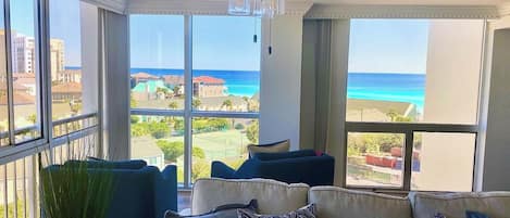 A room with a ceiling fan, large windows with a view of the ocean, a sofa with patterned cushions, and a potted plant.
