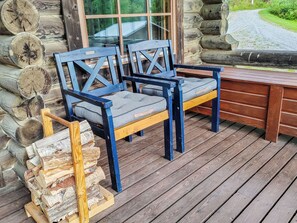 Une terrasse en bois avec deux chaises et une table, entourée de troncs d’arbres et donnant sur une fenêtre.