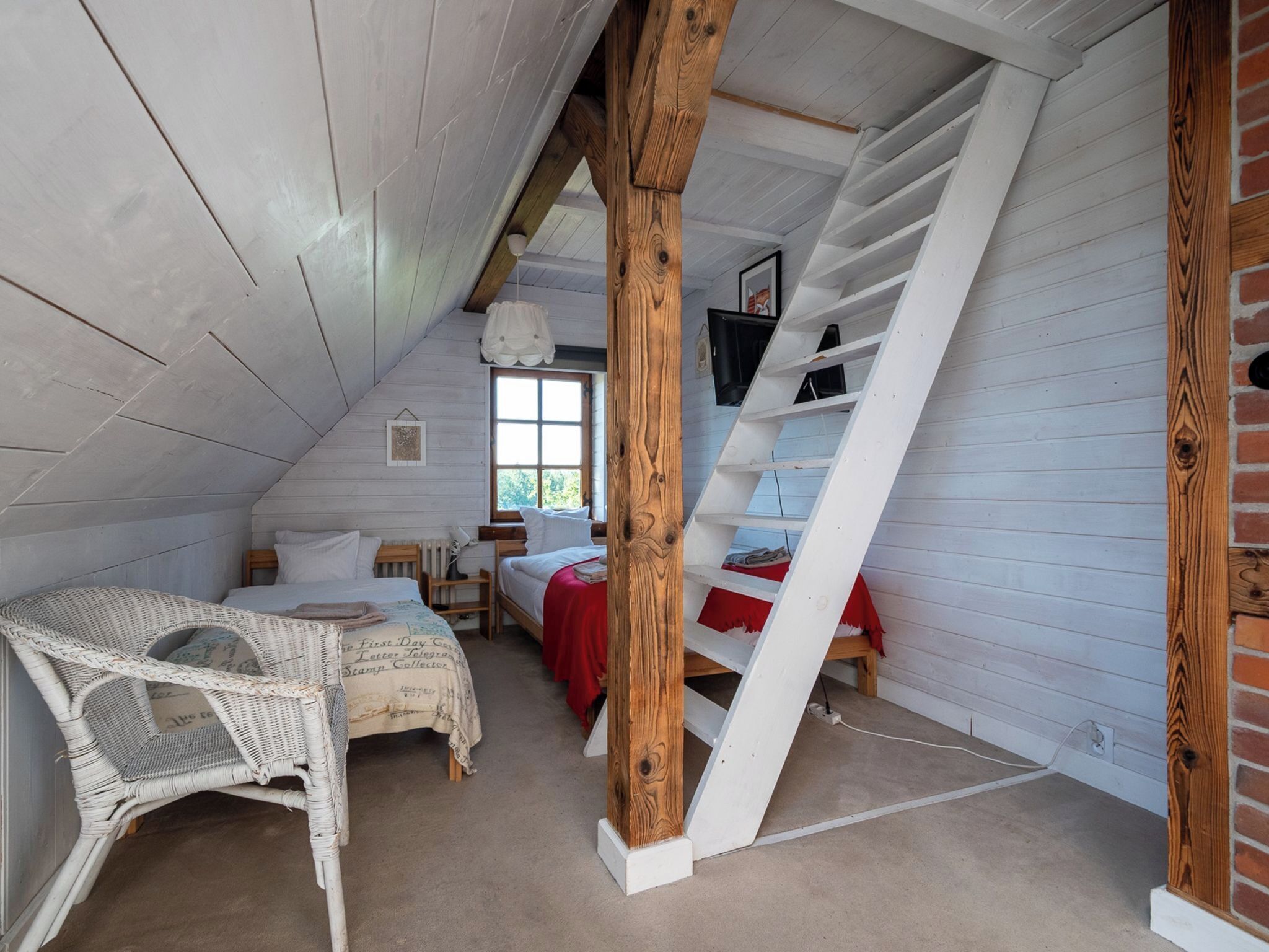 Een gezellige slaapkamer onder de dakrand, met schuine wanden, houten balken, een bed met een rode sprei, een rieten stoel en een raam.