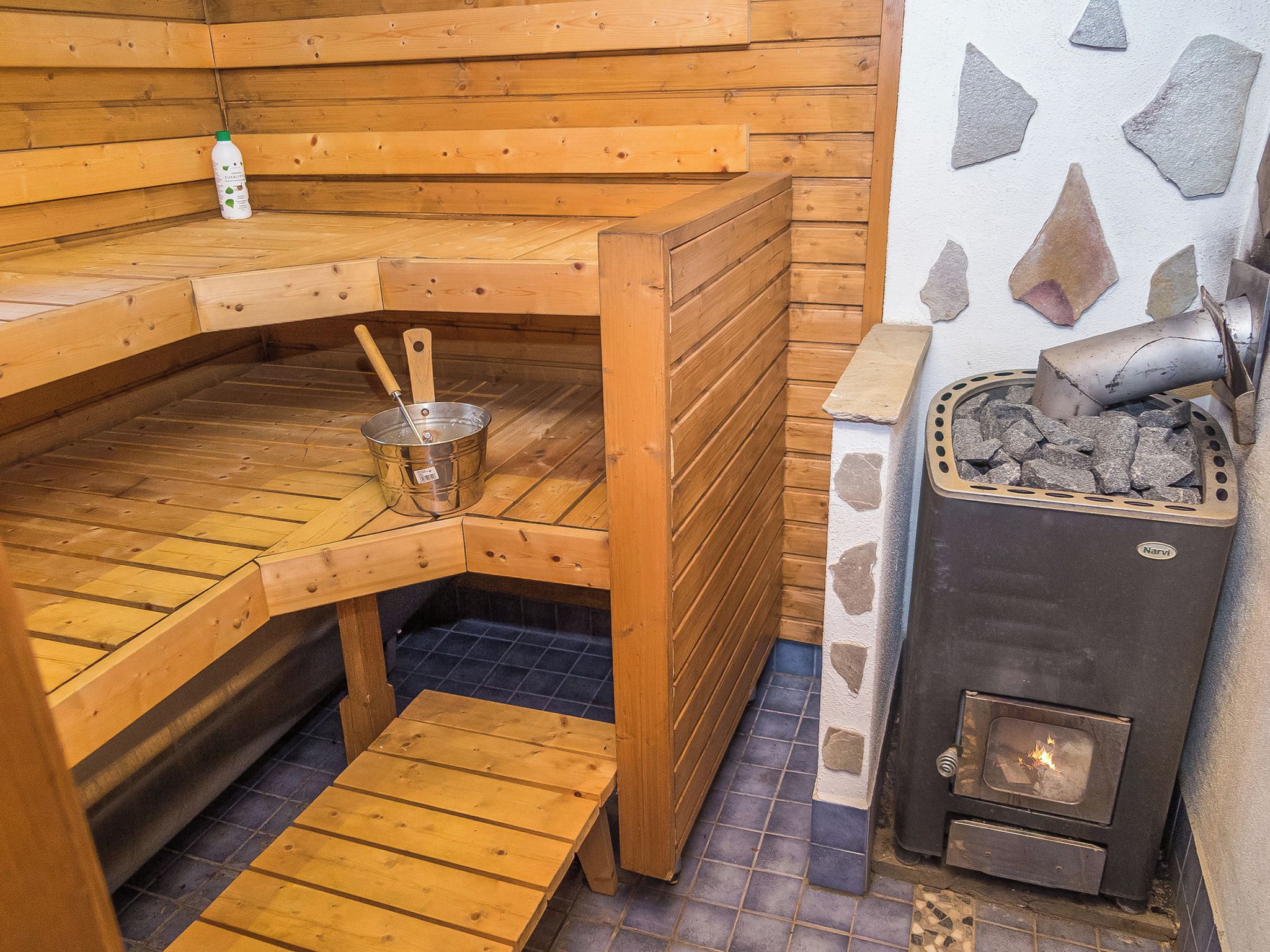 A sauna with wooden benches, a metal bucket, and a stone wall.