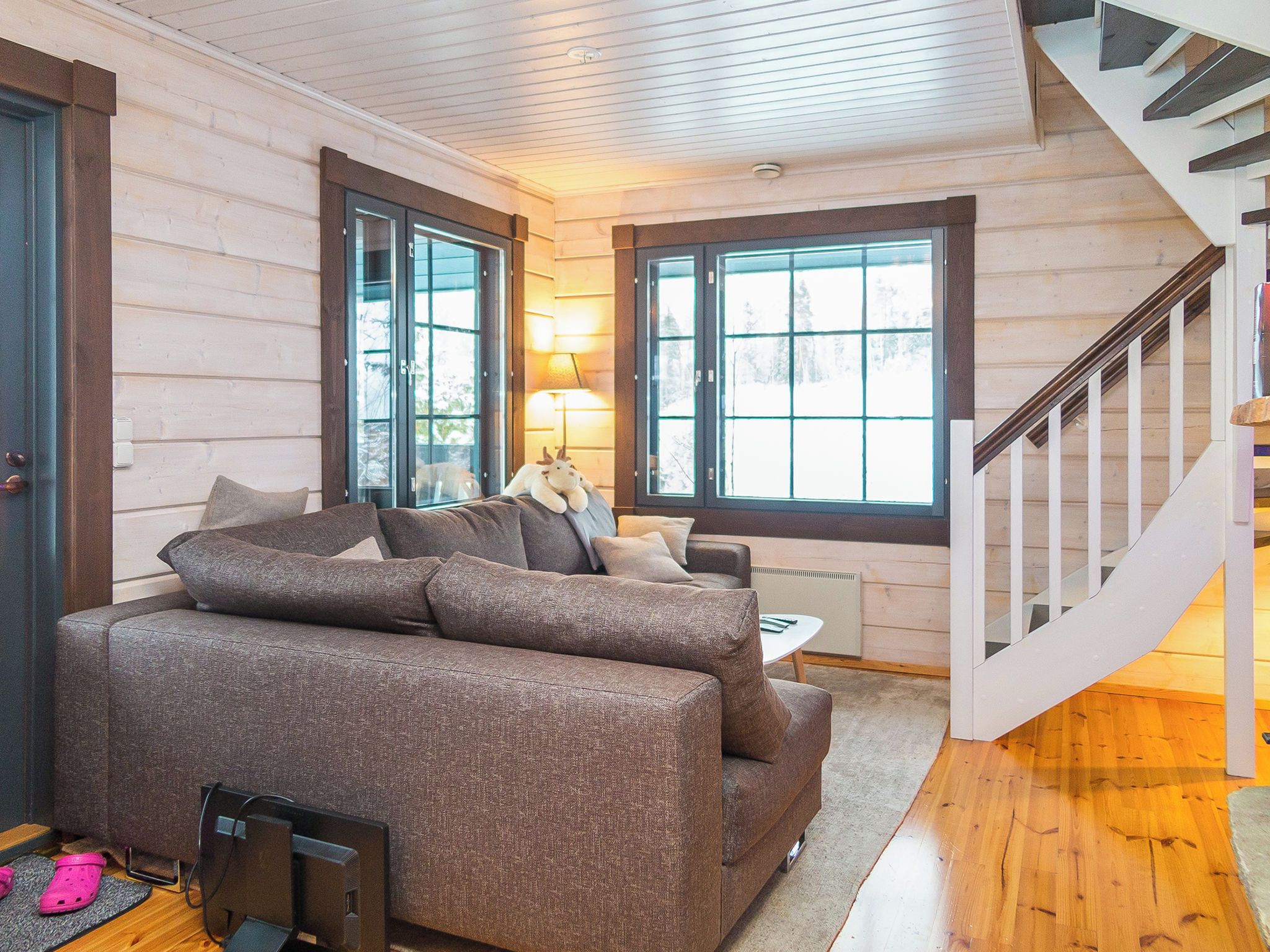 A cozy living room with a brown sofa, a lamp, and a view of the outside through large windows.