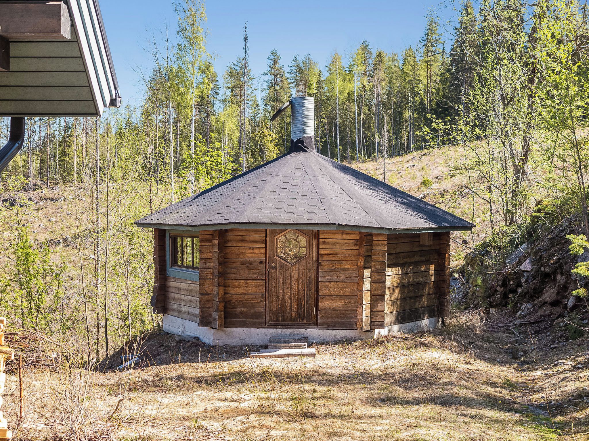 Eine Holzhütte mit einer sechseckigen Tür und einem Schornstein, umgeben von bewaldeter Landschaft.
